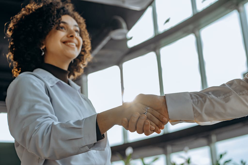 A smiling woman with curly hair shaking hands in a bright, modern office environment.